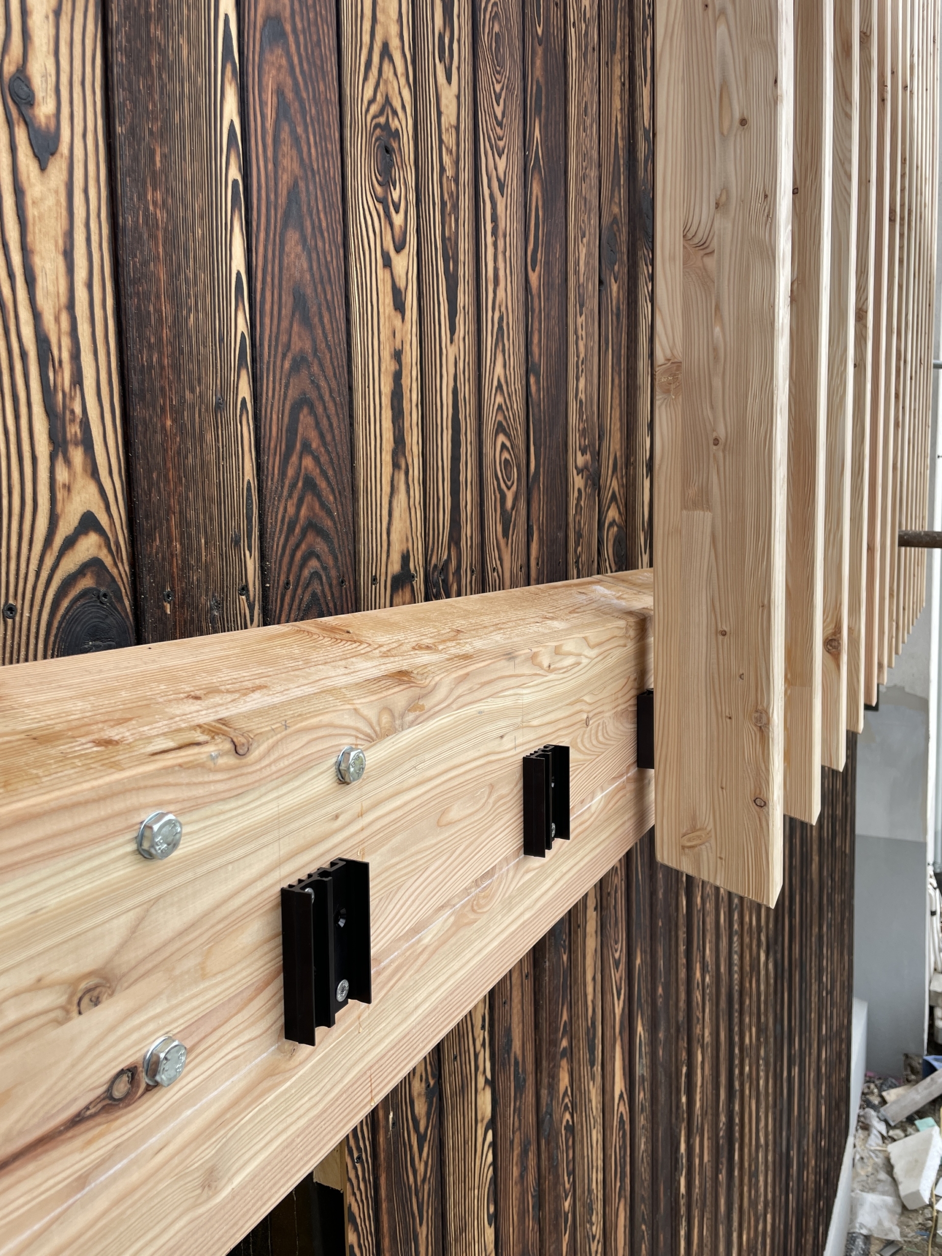 Houten gevel met geblakerde verticale planken en een horizontale balk met LamellenFix beugels en lamellen.